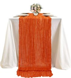 an orange table runner on top of a white table cloth with flowers in the center