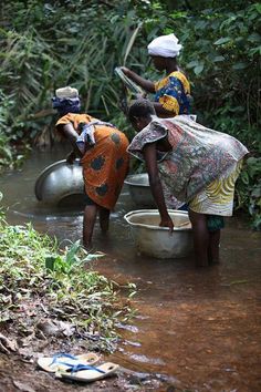 two women are washing their clothes in a small stream near some trees and bushes, while another woman is standing next to her