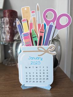 a desk calendar with scissors, markers and pencils in a jar on top of a wooden table