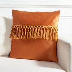 an orange pillow with tassels on it sitting on a white couch in front of a wall