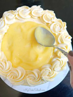 a person holding a spoon over a cake with white frosting and roses on it