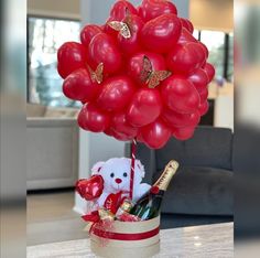 a teddy bear in a basket with red balloons and wine bottles on a table next to a couch