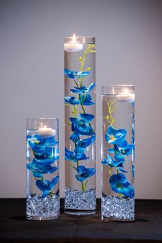 three tall glass vases with blue flowers and lit candles in them on a table