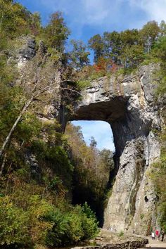 people are walking on the side of a river near a large rock formation with an opening in it