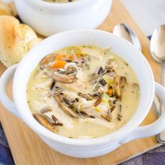 a bowl of chicken and wild rice soup on a cutting board with bread in the background