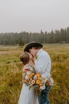 a bride and groom kissing in the middle of a field surrounded by tall grass with pine trees behind them