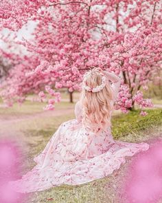 a woman in a pink dress sitting on the ground under a tree filled with pink flowers