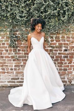 a woman standing in front of a brick wall wearing a white wedding dress with spaghetti straps