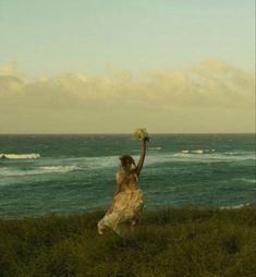 a woman in a white dress is walking by the ocean with her arms in the air