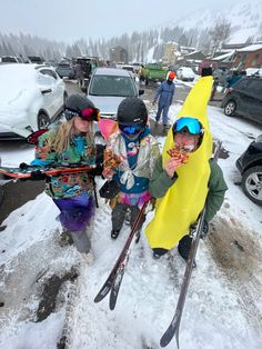 three children with skis and snowboards on a snowy road in front of parked cars