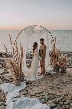 a bride and groom standing in front of an arch on the beach with their wedding dress draped over them