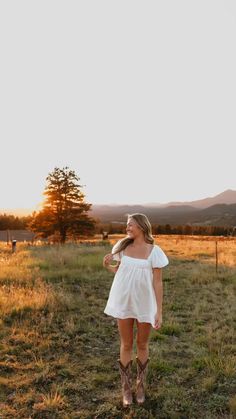 a woman in a white dress and cowboy boots is walking through an open field at sunset