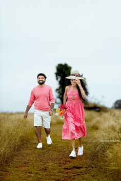 a man and woman walking down a dirt road holding hands while wearing pink dresses, hats and white shoes