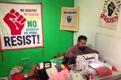 a man sitting at a sewing machine in front of a green wall with posters on it