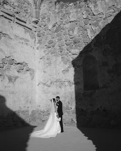 a bride and groom standing in front of a stone wall with their shadow on the ground