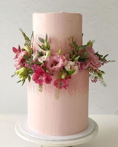 a pink frosted cake with flowers and greenery on the top is sitting on a white pedestal