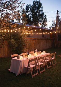 an outdoor dinner table set up in the backyard with string lights strung over it and bottles of wine on the table