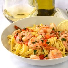 a white bowl filled with pasta and shrimp on top of a table next to a glass of wine