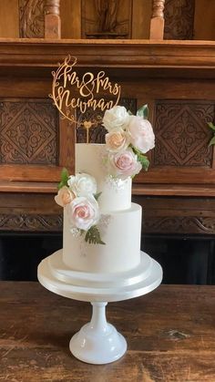 a white wedding cake with pink flowers on top is sitting on a table in front of a fireplace