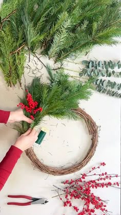 a woman is making a wreath out of branches and pine cones with red berries on it