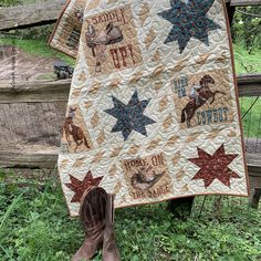 a cowboy themed quilt hanging from a wooden fence in the grass with boots and booties next to it