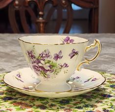 a tea cup and saucer with purple flowers on it sitting on a floral tablecloth