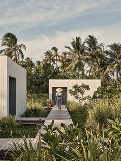 a woman walking down a wooden walkway between two white buildings with palm trees in the background