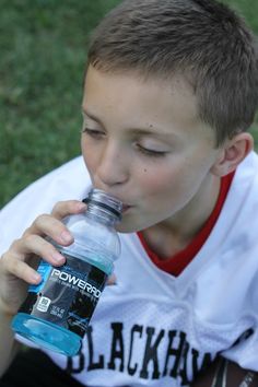 a young boy sitting on the grass drinking from a water bottle while wearing a white shirt