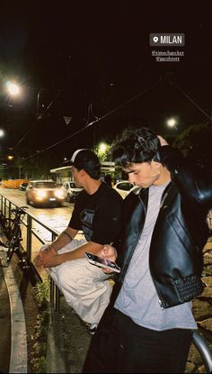 two young men standing next to each other on a sidewalk at night with their cell phones in their hands