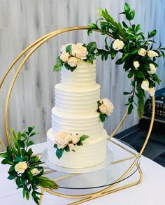 a white wedding cake with greenery and flowers on a round gold stand in front of a wall