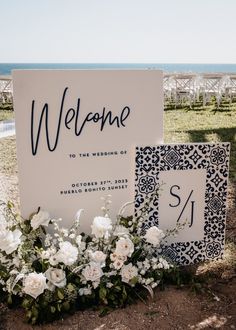 a welcome sign with white flowers and greenery in front of an ocean side ceremony