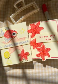 two cards with fish and flowers on them sitting on a bed next to a pen