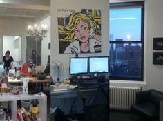 a woman is sitting at a desk in front of two computer monitors and a painting on the wall