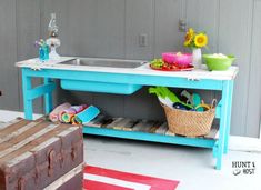 a blue table with baskets and bowls on it in front of a gray wall next to a wooden trunk