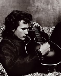 a black and white photo of a man holding a guitar in his right hand while sitting on a couch