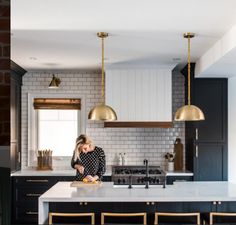 a woman standing in a kitchen with black cabinets and gold pendant lights hanging from the ceiling