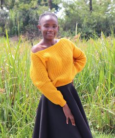 a woman standing in tall grass wearing a yellow sweater and black skirt with her hands on her hips