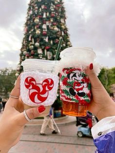 two people holding up cups with mickey and minnie mouse decorations on them in front of a christmas tree