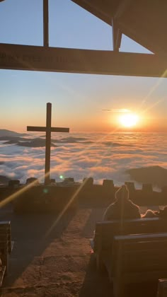 two people sitting on benches watching the sun rise over some clouds in the distance with a cross above them