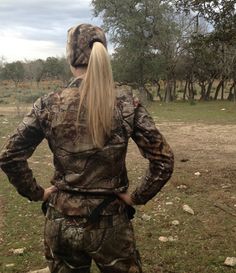 a woman with long blonde hair standing in the middle of a field wearing camouflage clothing