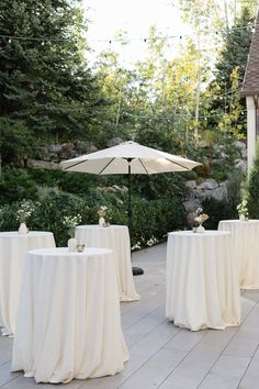 an outdoor dining area with tables covered in white cloths and umbrella, surrounded by greenery