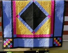 a quilt hanging on the side of a building in front of a planter filled with flowers