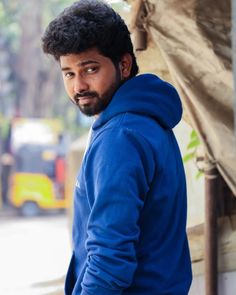 a man in a blue hoodie leaning against a wall and looking at the camera