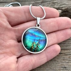 Our newest edition Northern Lights "Tree Tops" pendant - Large. Vibrant colors of Teal Greens, Blues, and Purples that DANCE in the light. We add a bit Copper and sparkle to represent the Milky Way and the Stars. Our original mixed media hand painted artist rendition of the Northern Lights Hand crafted in Upper Michigan 2 Large statement shapes round or rectangle smaller petite charm sizes also available 18" Stainless Snake chain with 2" extender. Other chain options available message us, includ Upper Michigan, The Milky Way, The Northern Lights, Tree Tops, Tree Lighting, Copper Pendants, Wrap Rings, Round Pendant, Stainless Steel Jewelry