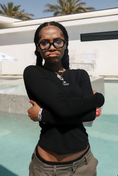 a woman with her arms crossed standing in front of a swimming pool wearing black glasses