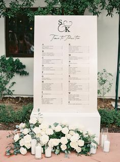 wedding seating chart with white flowers and candles on the ground next to a large sign