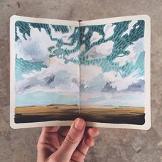 a hand holding up an open book with watercolors on the pages and trees in the background