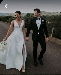 a bride and groom are walking down the road holding hands with each other, dressed in black tuxedos