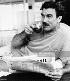 a man with a moustache drinking from a glass while reading the news paper