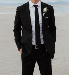 a man in a suit and tie standing on the beach with his hands in his pockets